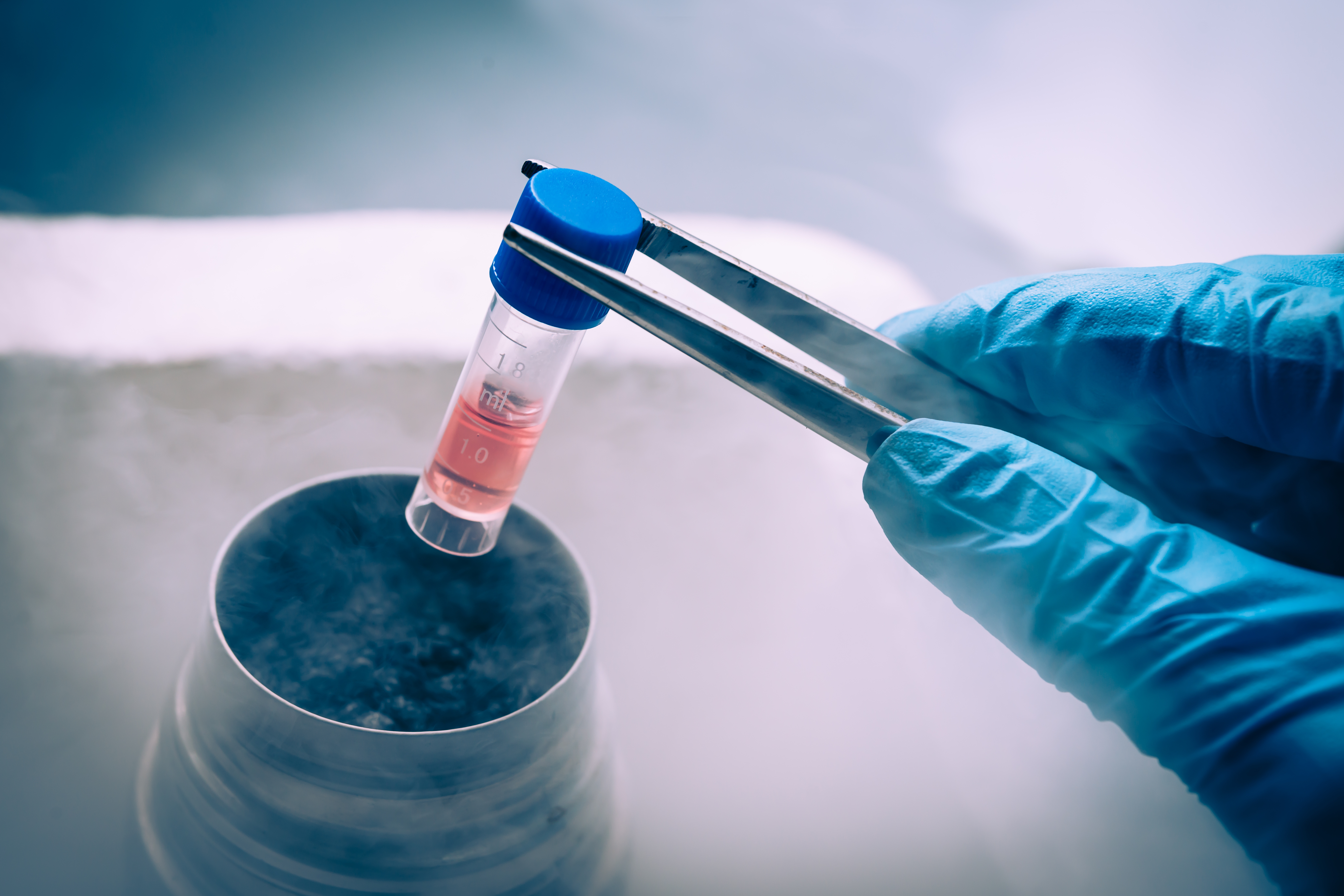 A gloved hand uses forceps to place a vial of liquid into a thermal container surrounded by fog.